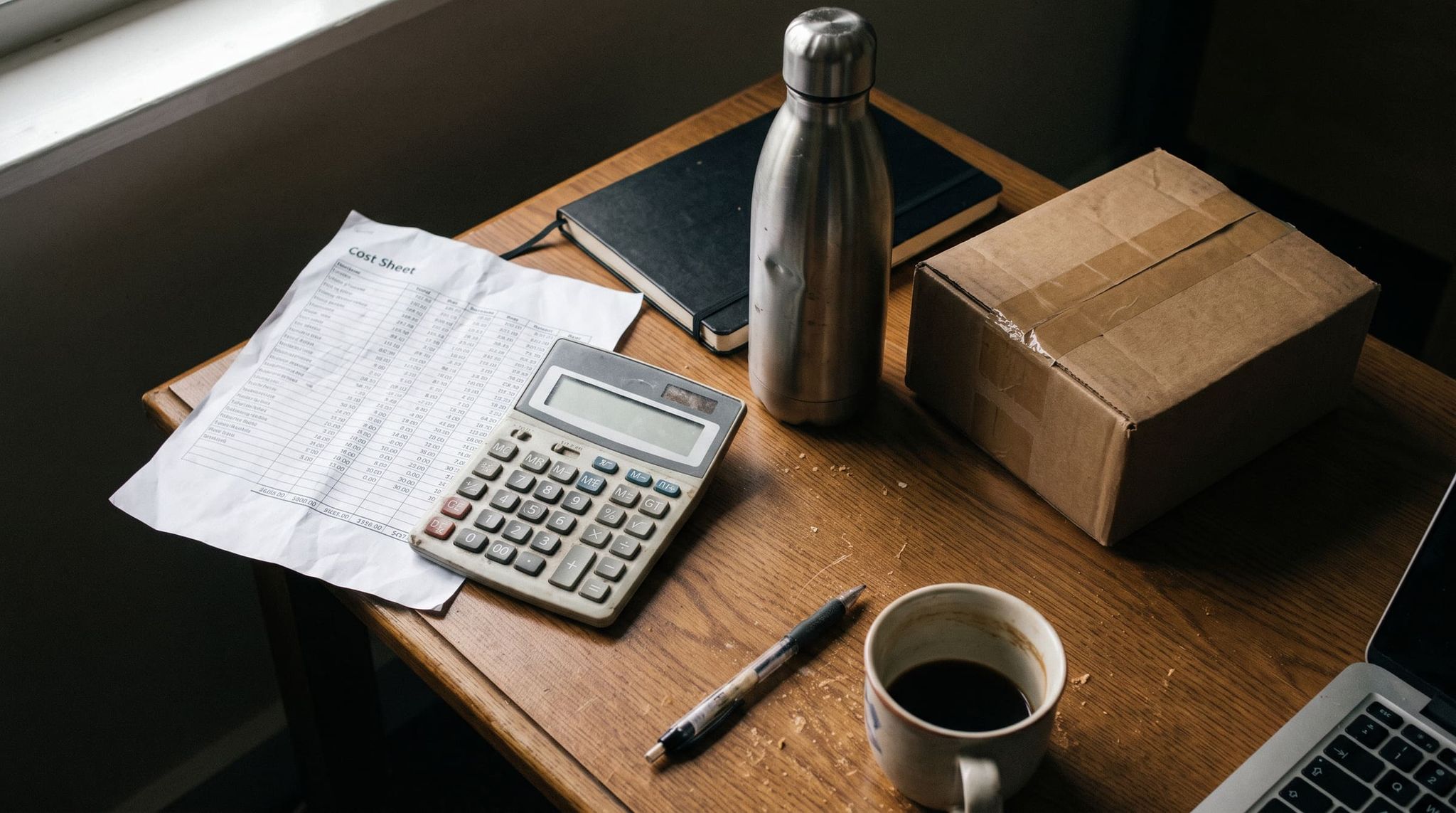 Printed cost sheet, calculator, stainless steel insulated bottle, shipping carton and coffee mug on a wooden desk