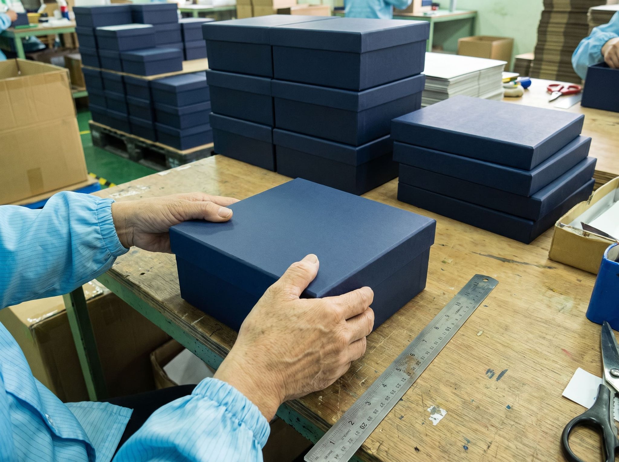 Two hands adjusting the lid of a navy blue rigid gift box on a workbench, steel ruler and stacks of boxes