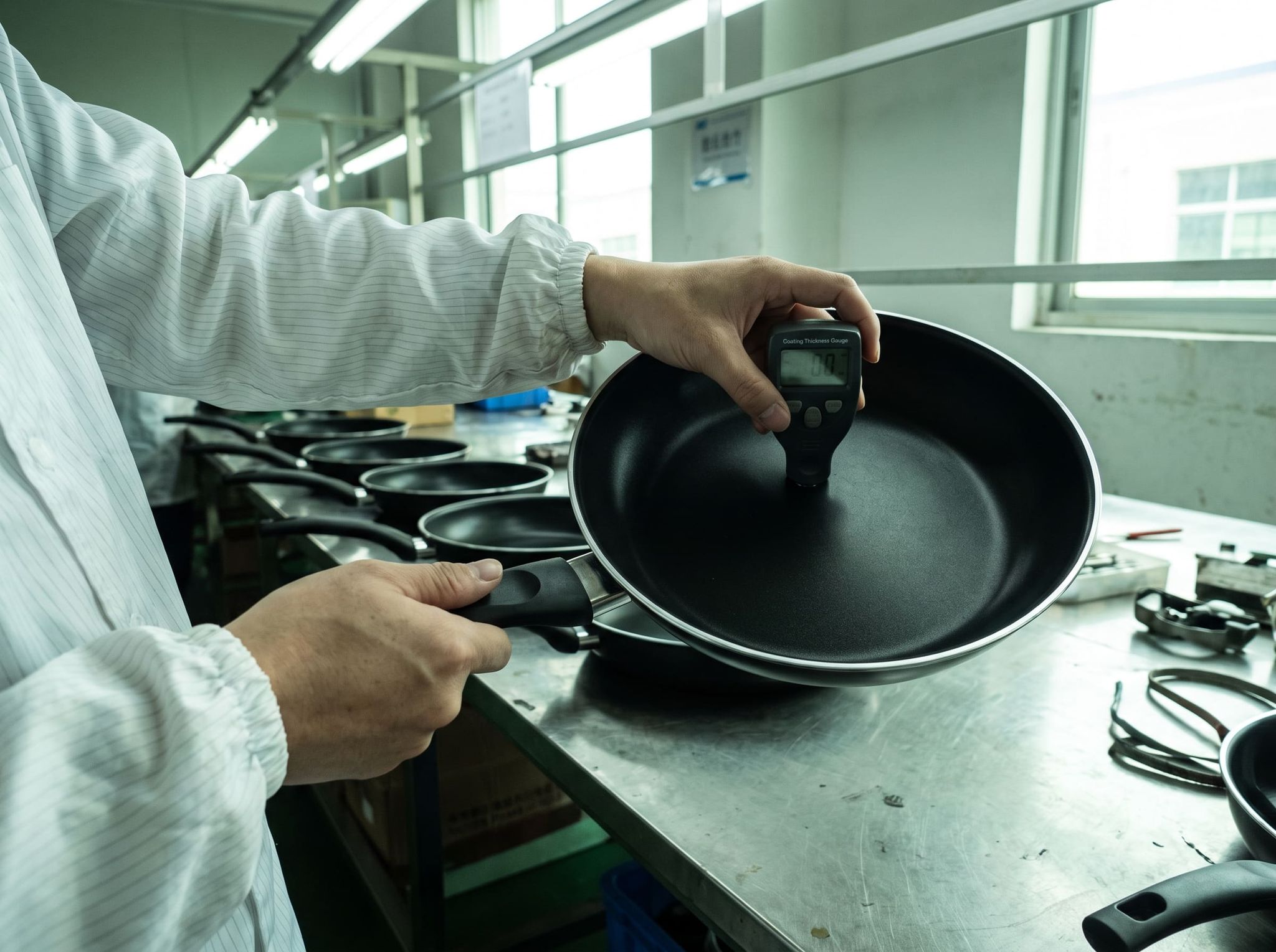 Measuring the thickness of a pan's non-stick coating with a gauge, row of pans on the workbench