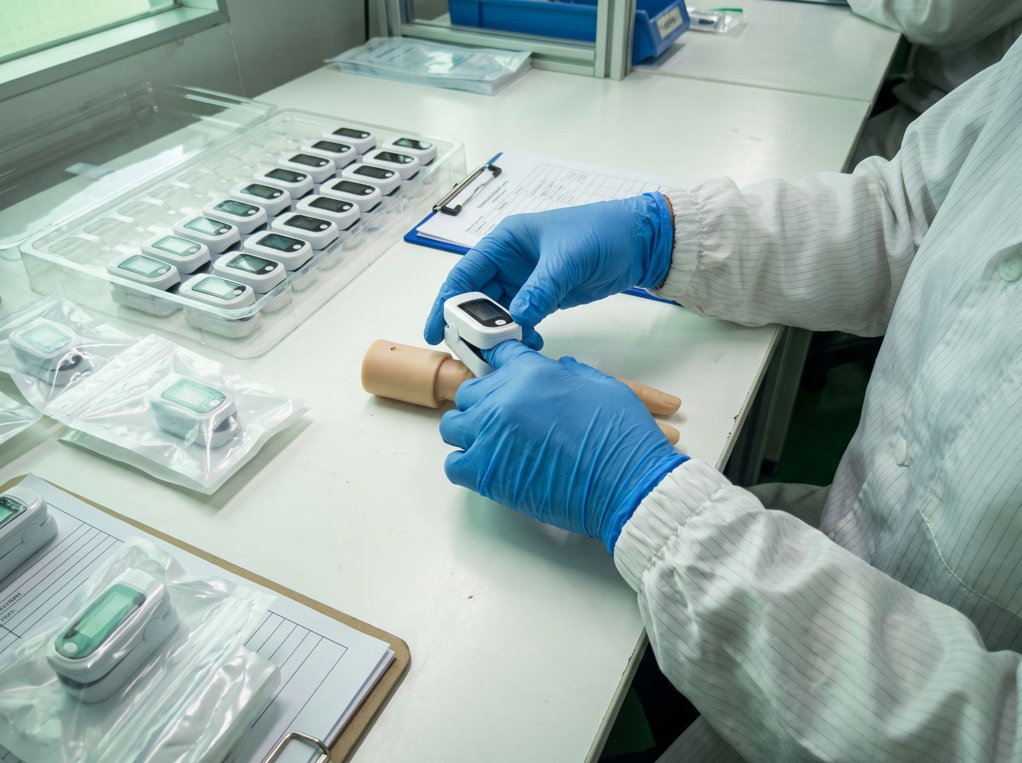 Hands in blue nitrile gloves testing a pulse oximeter, near a tray of identical devices and sealed pouches