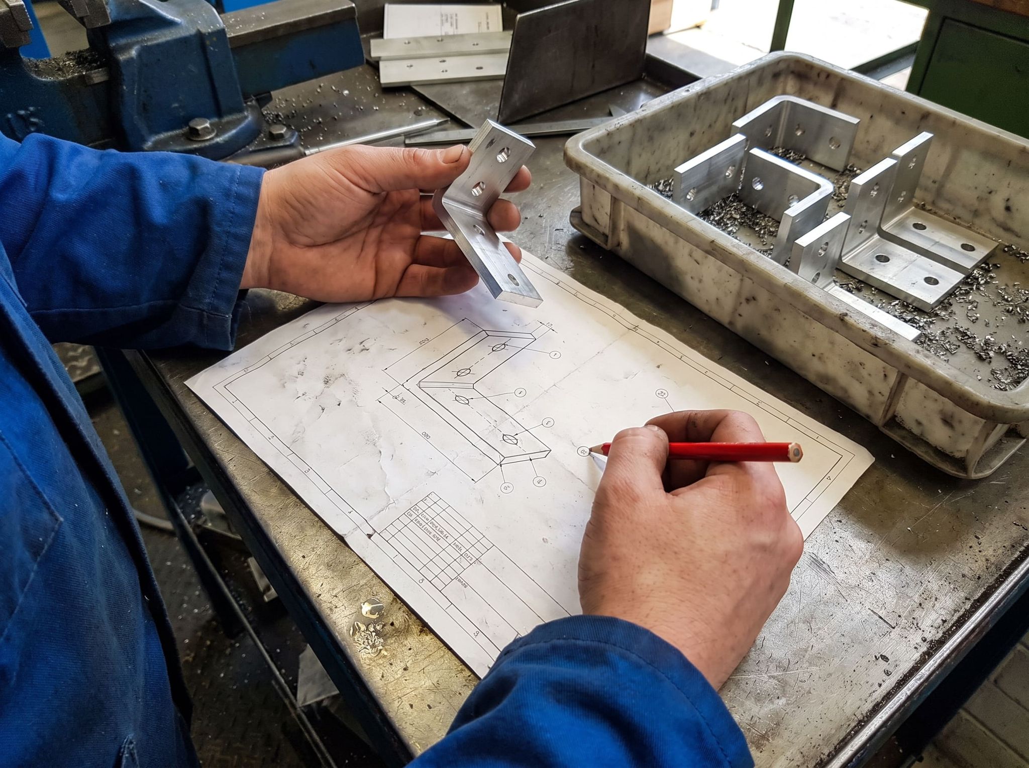 Machined part compared with its drawing: one hand holds the aluminum square, the other ticks a dimension with a red pencil