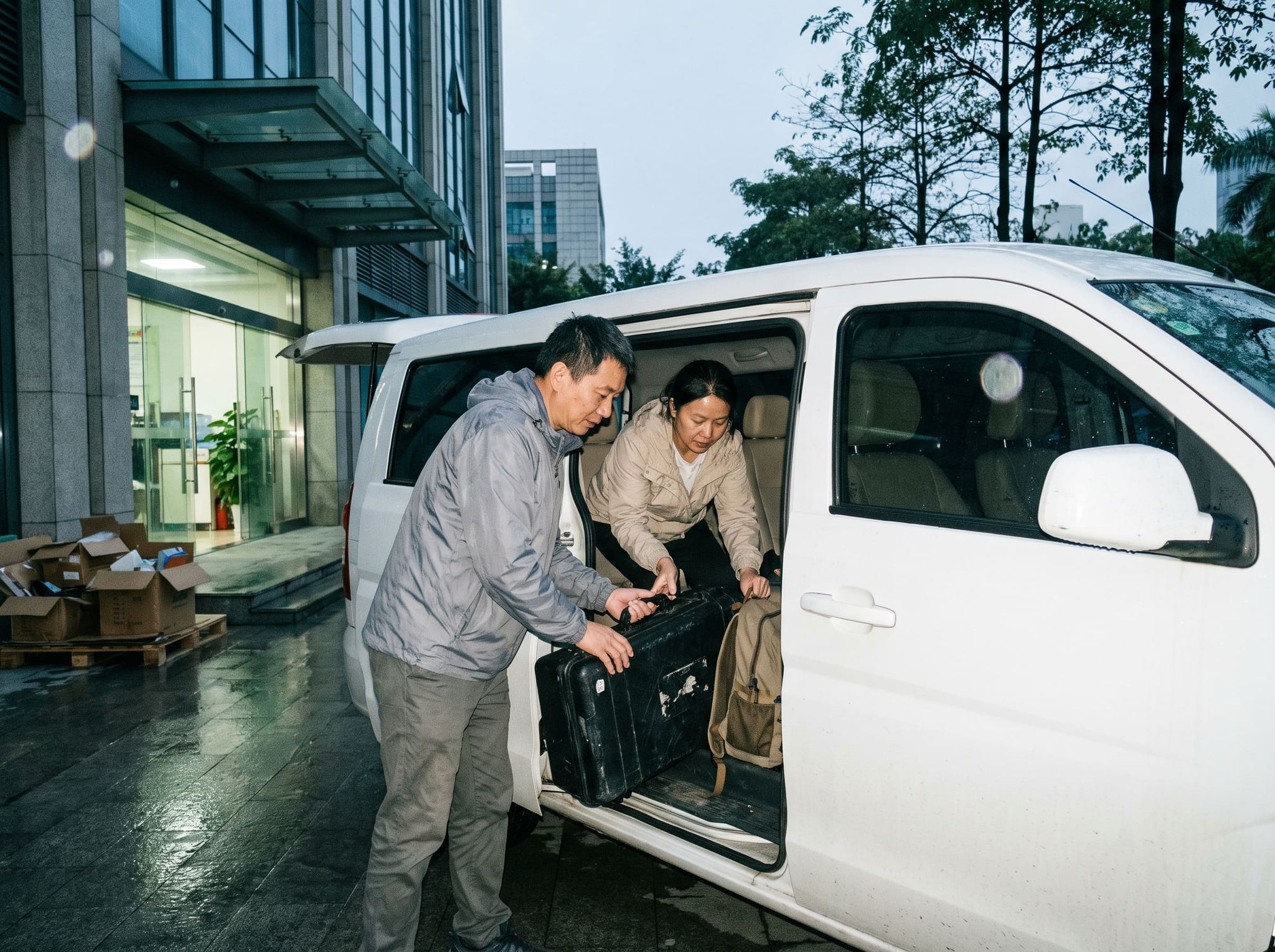 At dawn, two people load a case of equipment and a backpack into a white minivan in front of an office building