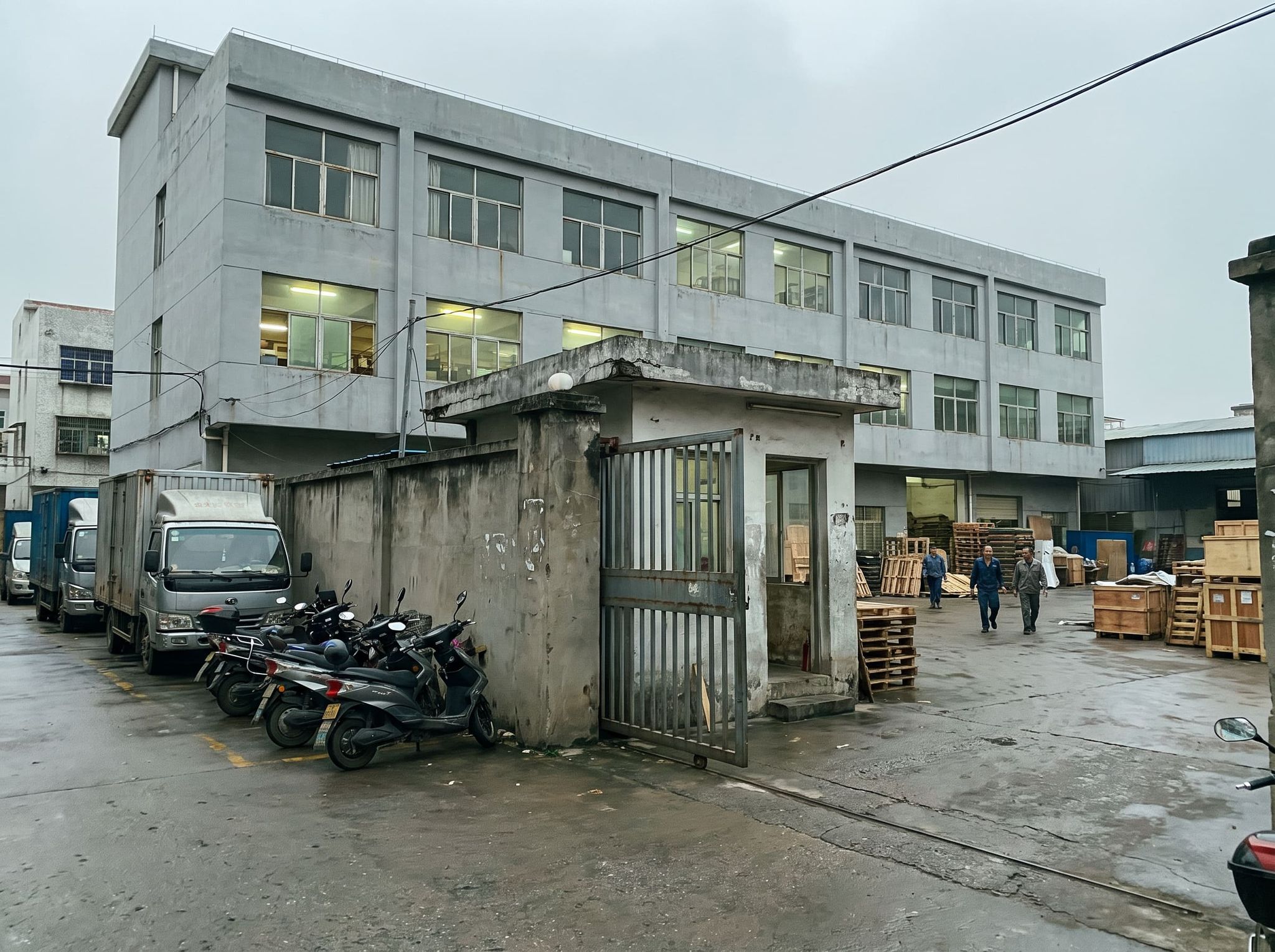 Entrée d'usine par temps couvert : portail en acier, guérite, bâtiment gris de trois niveaux, camions, scooters, trois ouvriers dans la cour