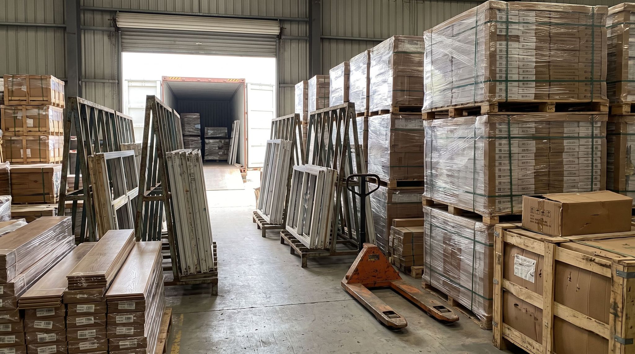 Materials export warehouse: wrapped pallets, window frames on trestles, flooring planks and a pallet jack in front of a container