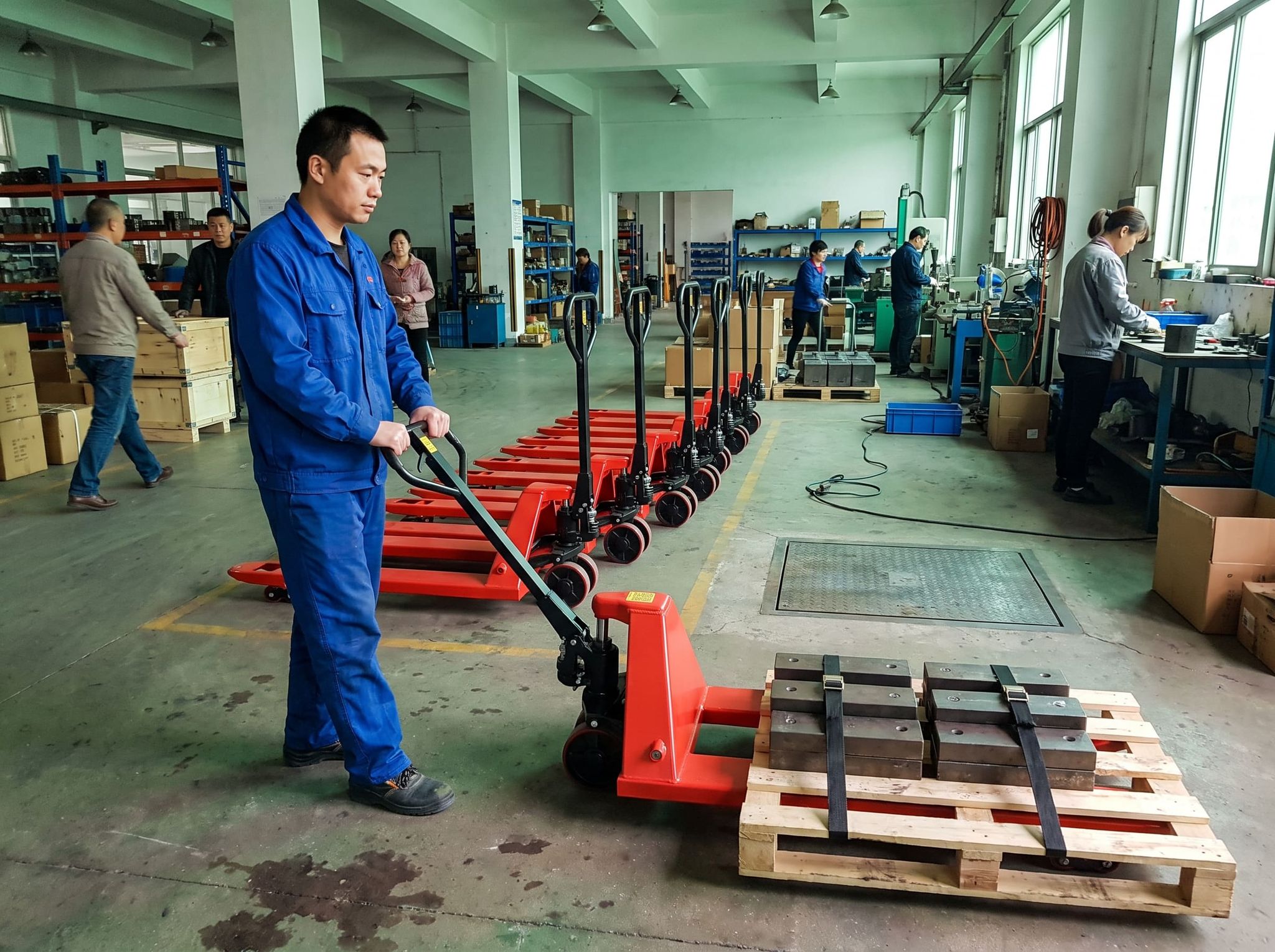 Technician in blue coveralls operating a red manual pallet jack loaded with strapped test weights in a workshop