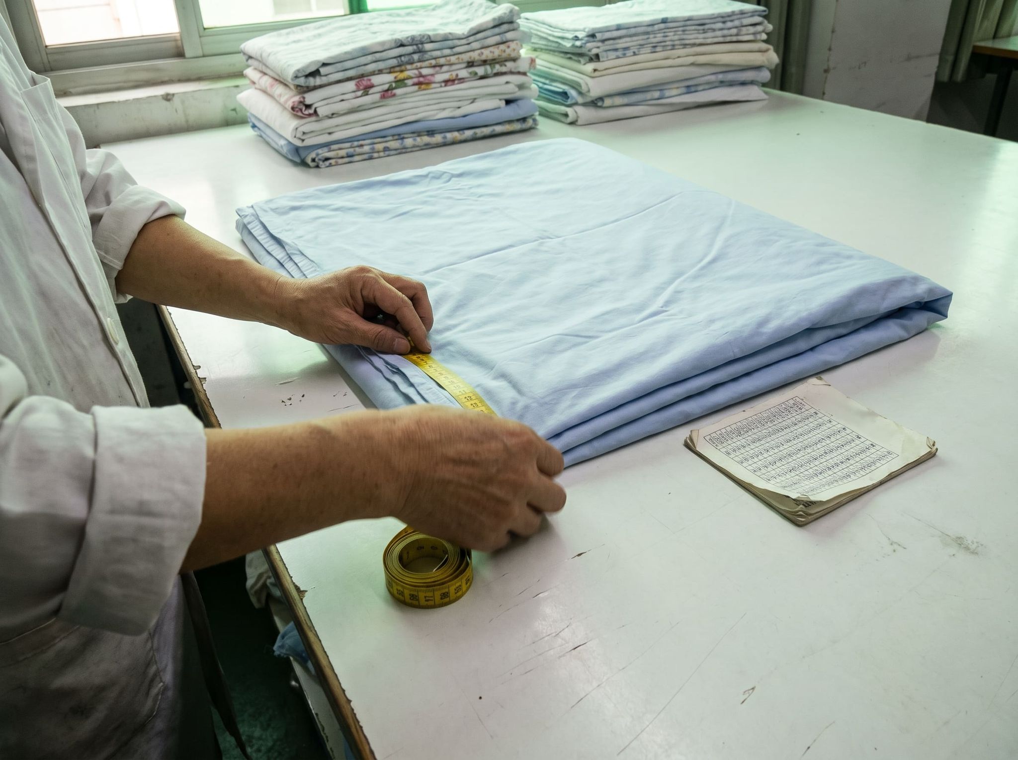 Two hands measuring with a tape the edge of a pale blue duvet cover folded on an inspection table