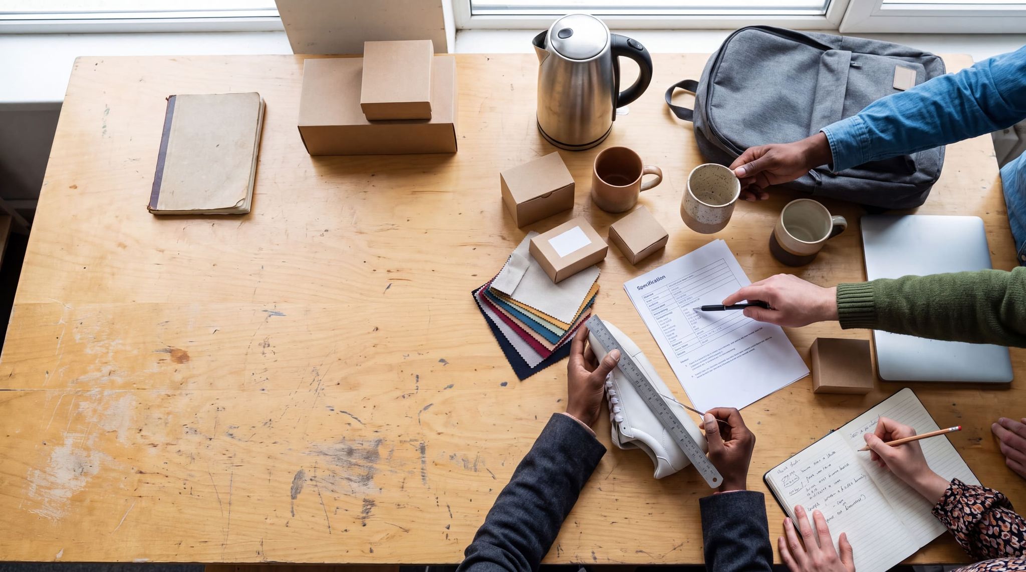 Top view of a sample table: several hands examining a mug, a sneaker, a spec sheet and color swatches