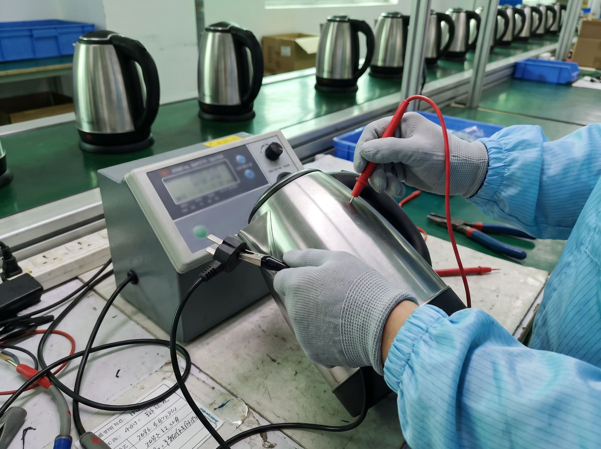 Gloved hands testing the electrical safety of a stainless steel kettle with a test probe, on an end-of-line test bench