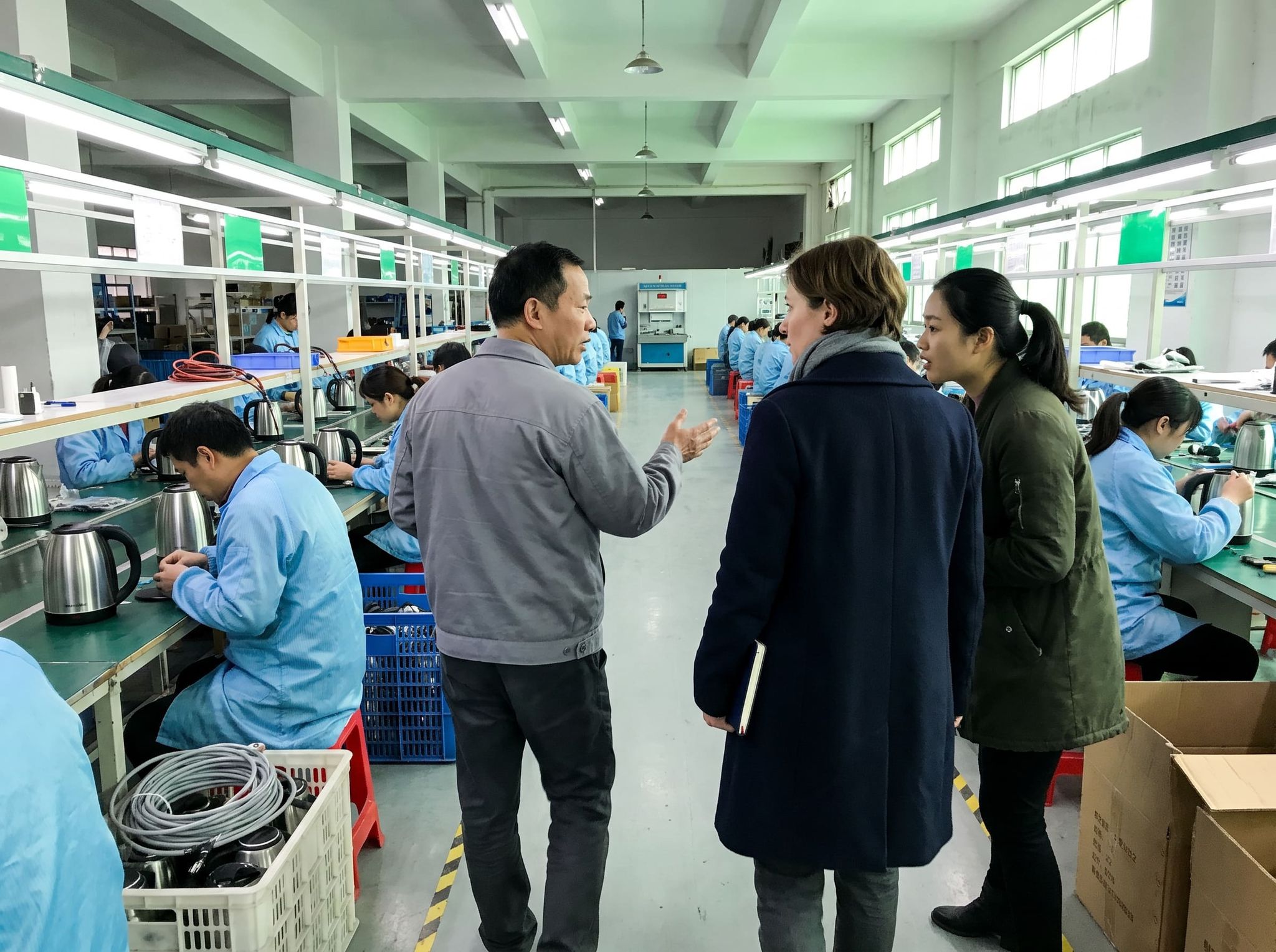 Factory visit: in the aisle of a kettle assembly line, the production manager explains the process to a buyer, accompanied by her interpreter