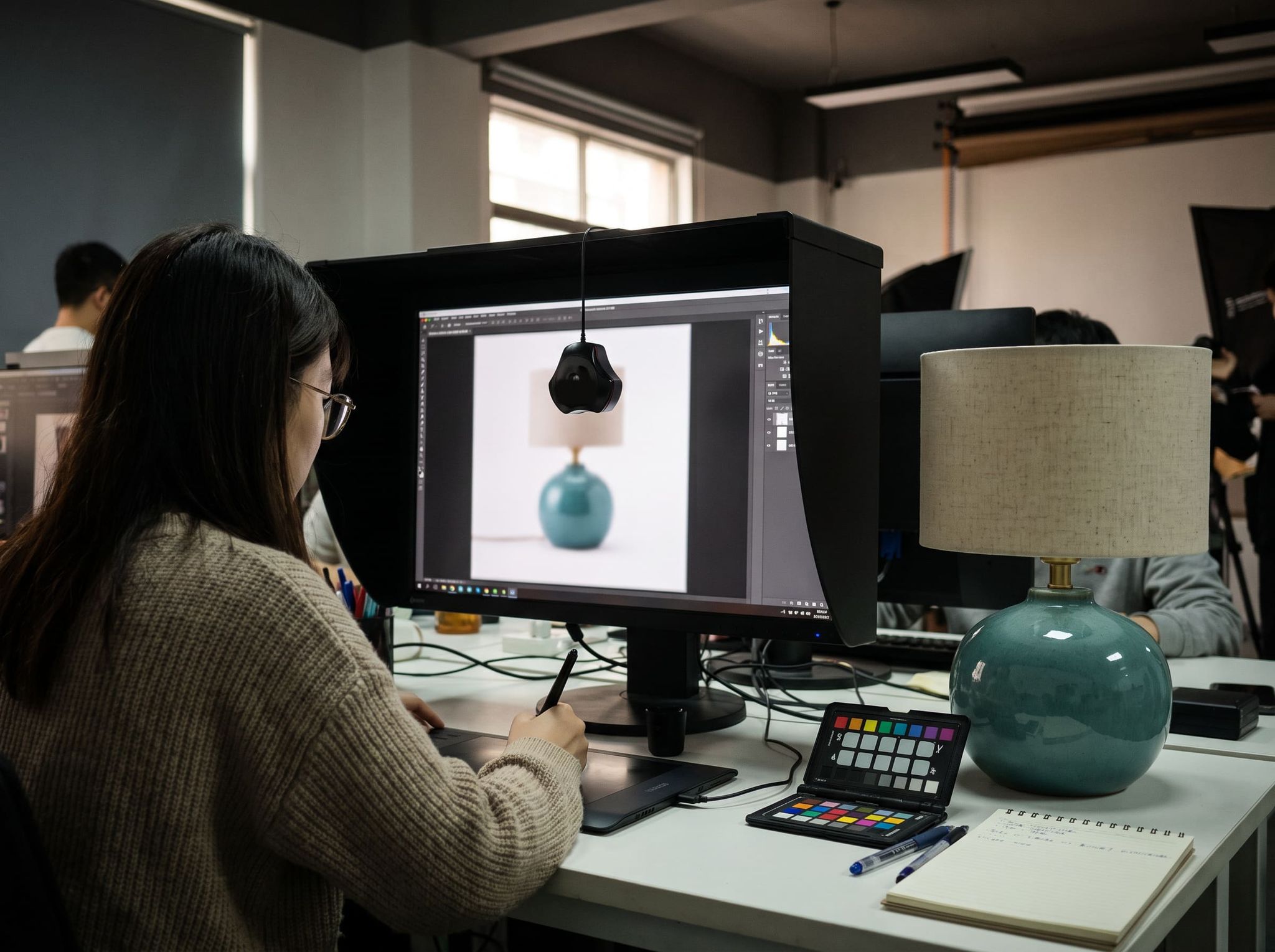 Retoucher working on a graphics tablet in front of a probe-calibrated screen, the reference ceramic lamp switched off beside it, color chart on the desk