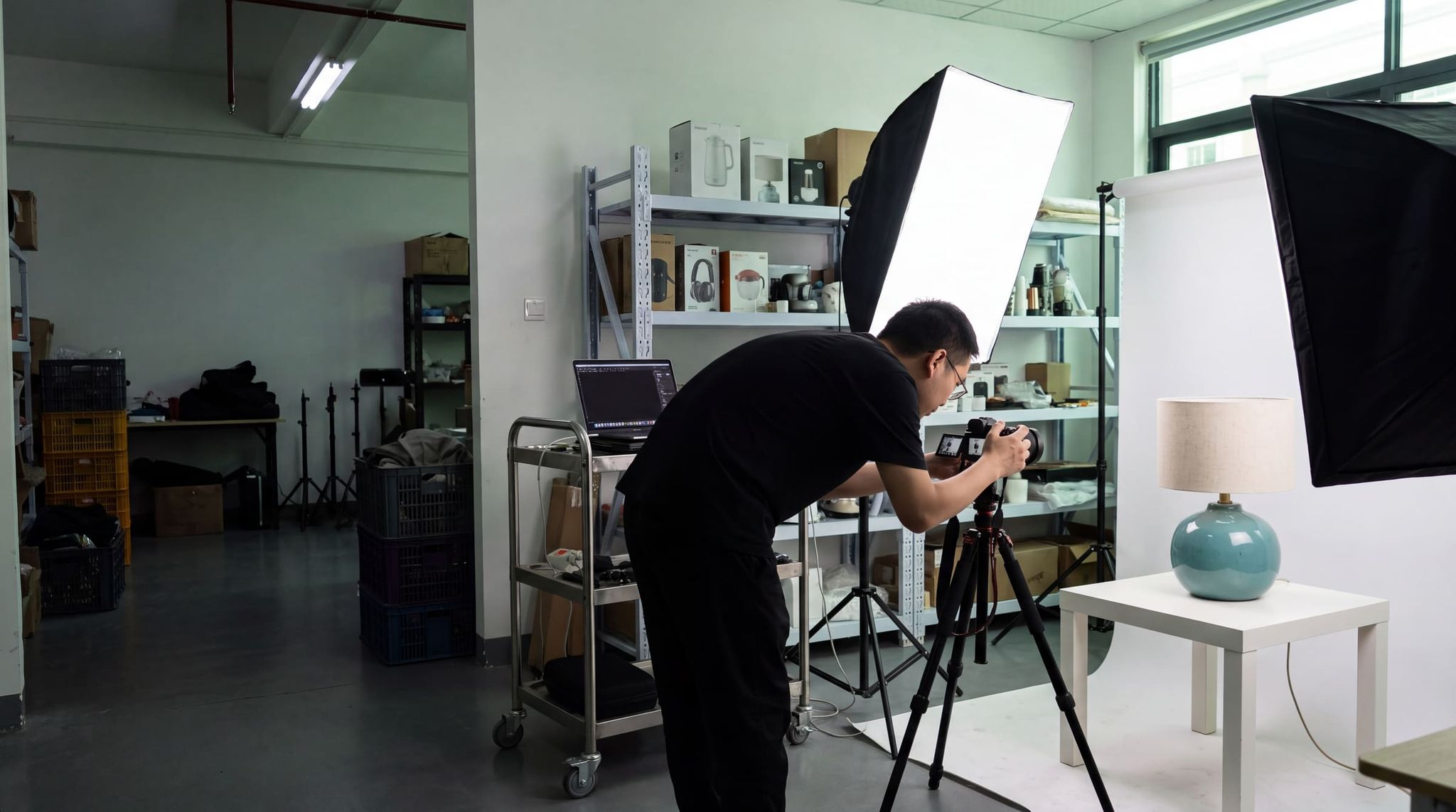 Photographer in a black T-shirt adjusting his camera on a tripod facing a blue-green ceramic lamp, between two softboxes